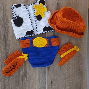 May include: A crochet baby outfit resembling a cowboy costume. The set includes a white vest with black cow spots, an orange cowboy hat, blue diaper cover with an orange belt, orange chaps with "Andy" embroidered, and orange booties.
