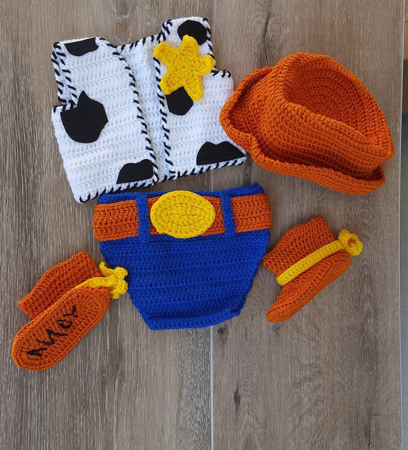 May include: A crochet baby outfit resembling a cowboy costume. The set includes a white vest with black cow spots, an orange cowboy hat, blue diaper cover with an orange belt, orange chaps with "Andy" embroidered, and orange booties.