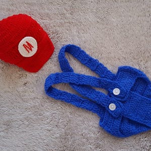 May include: A red crocheted hat with a white circle and a red "M" and a blue crocheted overall set with white buttons. The set is designed for a baby or toddler and is likely a costume.