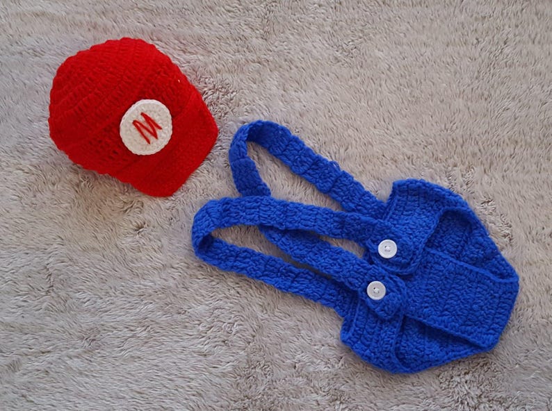 May include: A red crocheted hat with a white circle and a red "M" and a blue crocheted overall set with white buttons. The set is designed for a baby or toddler and is likely a costume.