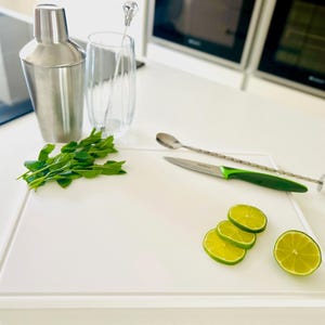 May include: A cocktail-making setup featuring a stainless steel shaker, a tall glass, and fresh mint. A white chopping board holds sliced limes, a knife with a green handle, and a bar spoon, all ready for preparing cocktails.