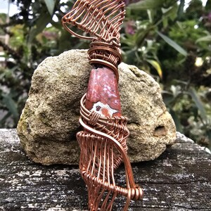 May include: A handcrafted pendant featuring a red and white stone wrapped in copper wire. The wire is intricately woven around the stone, creating a cage-like design. The pendant is displayed on a weathered wooden surface, with a blurred natural background.