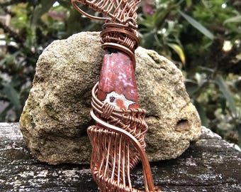 May include: A handmade pendant with a red and white stone encased in copper wire. The wire is carefully woven around the stone, forming a cage-like structure. The pendant is shown on a weathered wooden surface, with a blurred natural backdrop.