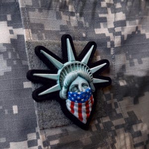 May include: A black and white patch of the Statue of Liberty wearing a red, white, and blue bandana over her face. The patch is attached to a camouflage fabric.