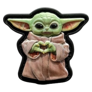 May include: A green and tan character from the Star Wars franchise, making a heart shape with its hands. The character is wearing a tan robe and has large black eyes and large ears. The image has a black border.