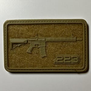 May include: A rectangular, olive drab patch featuring a detailed depiction of a rifle. The patch includes the text ".223" and the brand name "L-TAC". The patch has a hook and loop backing.