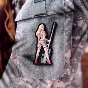 May include: A gray patch with a woman in a black and white outfit holding a rifle. The woman has blonde hair and is wearing red high heels. The patch is on a camouflage background.
