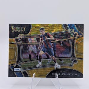 May include: A gold and blue trading card featuring a footballer in a red and blue kit. The card has the text "Select" and "FIFA" on it. The player is Robert Lewandowski, who plays for FC Barcelona.