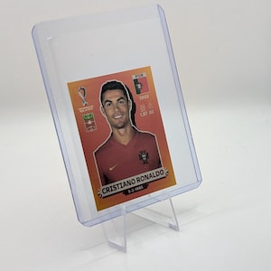 May include: A clear plastic card holder displaying a Cristiano Ronaldo trading card. The card features a portrait of Ronaldo in a red jersey, with the text "CRISTIANO RONALDO" and his birthdate. The card is supported by a clear plastic stand.