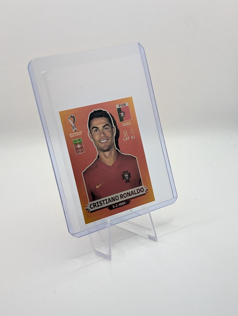 May include: A clear plastic card holder displaying a Cristiano Ronaldo trading card. The card features a portrait of Ronaldo in a red jersey, with the text "CRISTIANO RONALDO" and his birthdate. The card is supported by a clear plastic stand.