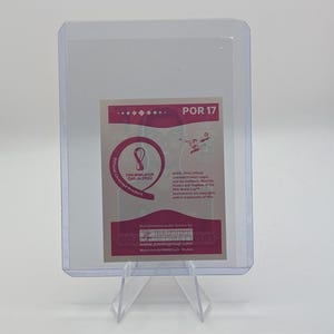 May include: A trading card featuring the FIFA World Cup Qatar 2022 logo and text, protected in a clear plastic holder. The card has a pink and white design with the number "17" visible.