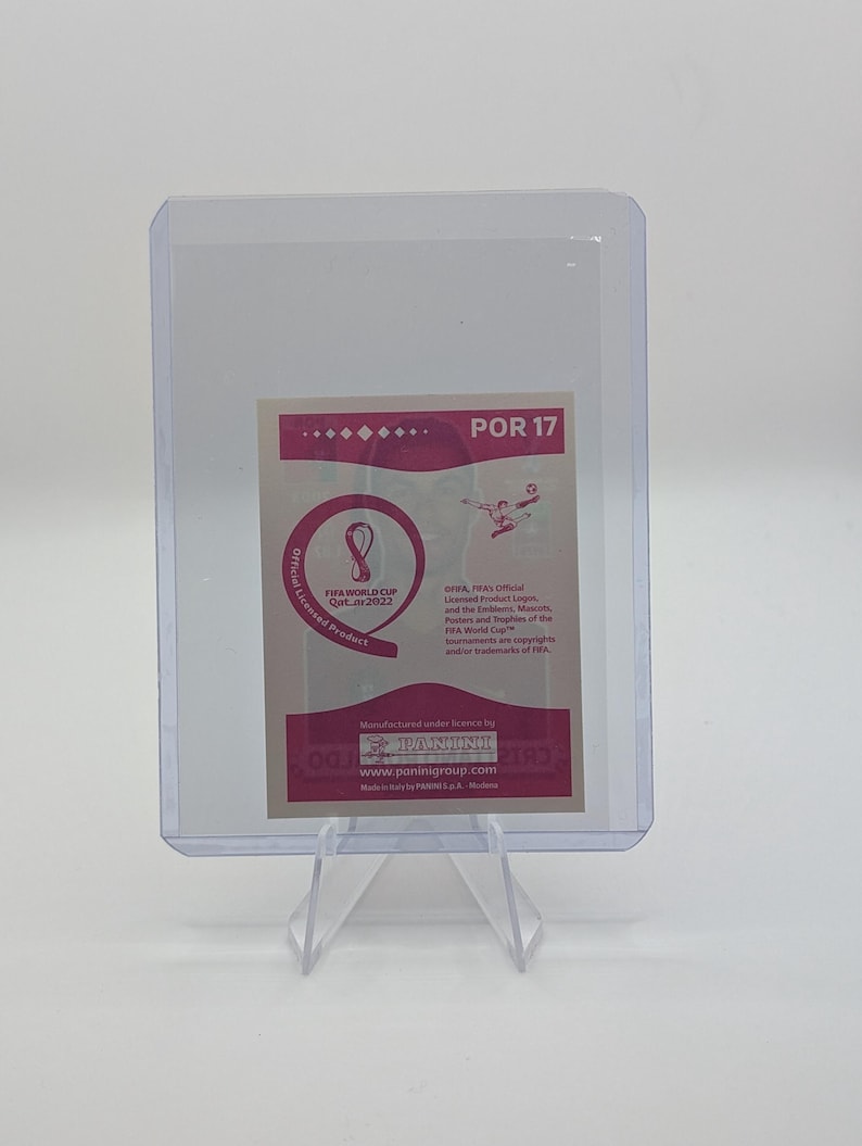 May include: A trading card featuring the FIFA World Cup Qatar 2022 logo and text, protected in a clear plastic holder. The card has a pink and white design with the number "17" visible.