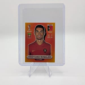 May include: A Cristiano Ronaldo trading card in a protective sleeve. The card features a portrait of Ronaldo in a red jersey, with the text "CRISTIANO RONALDO" and the date "5-2-1985". The card also includes the Portuguese flag and other details.