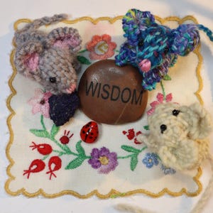 May include: A handcrafted scene featuring three knitted mice in gray, blue, and cream, arranged on an embroidered cloth with floral patterns. A stone with the word "WISDOM" is present, along with a red ladybug. The scene is a whimsical display.