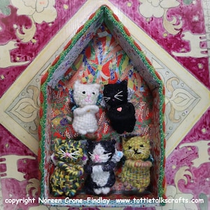 May include: A handcrafted wooden cat house with six small, crocheted cat figurines inside. The cats are white, black, yellow, and calico, each with embroidered features. The house is decorated with patterned paper and trim, set against a pink and cream background.