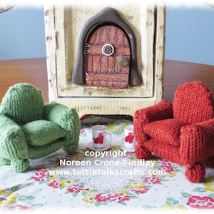 May include: Two knitted armchairs, one green and one red, are placed on a floral tablecloth in front of a miniature fireplace with a door. The text "copyright Noreen Crone-Findlay www.tottietalkcrafts.com" is visible below the chairs.
