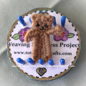 May include: A small, handmade brown teddy bear figure stands on a white circular base with blue pegs. The base has floral designs and text that reads "Weaving Project" and "tottiecrafts.com". The bear has black eyes and is made of yarn.