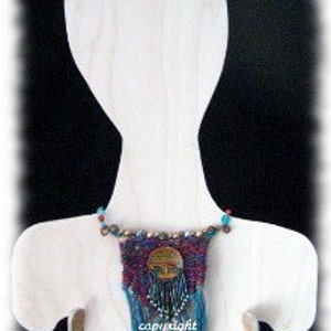 Puede incluir: Un collar de cuentas con un colgante de tapiz tejido que presenta un diseño circular con una cara y flecos. El tapiz está en tonos de morado, rojo y azul.