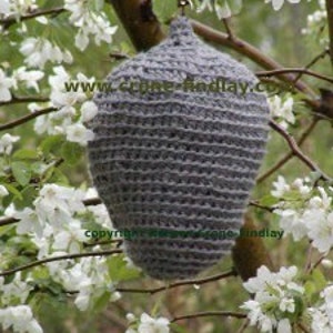 May include: A gray crocheted birdhouse hanging from a tree branch with white blossoms. The birdhouse is shaped like a teardrop and has a textured surface. The website www.crene-findlay.co.uk is visible on the birdhouse.