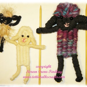 May include: Three yarn dolls, two black sheep and one white lamb, are standing on wooden sticks. The black sheep have purple, blue, and green yarn for their bodies. The white lamb has a pink nose and black eyes. The dolls are handmade and have a whimsical, folk art style.