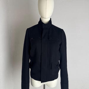 May include: A black jacket with a high collar, long sleeves, and a front button placket. The garment features two rectangular chest flaps with buttons and a structured fit, displayed on a mannequin against a plain, light-colored background.