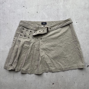 May include: A khaki-colored mini skirt featuring a pleated design on one side, a waist belt with a metal buckle, and a series of small metal studs along the front panel. The interior label displays the brand name MOXX in white text on a black background.