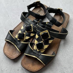 May include: Black leather sandals with gold accents and black gemstones. The sandals have a yellow and black geometric pattern on the straps. The sandals have the word "MIU MIU" written on the straps.