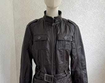 May include: A dark brown jacket with a high collar, a full-length front zipper, and a matching belt with a rectangular buckle. The jacket features two chest pockets with buttoned flaps, shoulder epaulets, and long sleeves with stitched panel details.