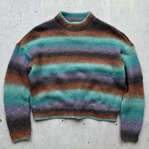 May include: A striped sweater in shades of brown, teal, purple, and gray. The sweater has a crew neck and long sleeves.