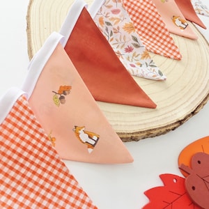 May include: A fabric bunting banner featuring triangular flags in various patterns including orange gingham, solid burnt orange, autumn leaves, and a light peach print with foxes and acorns, all attached to a white fabric header tape.