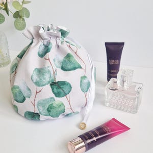 May include: A grey and white cosmetic bag with a green eucalyptus leaf design. The bag is next to two tubes of lotion, a perfume bottle, and a sprig of greenery. The bag is tied with a white ribbon and a gold bead. The bag is ideal for storing toiletries.