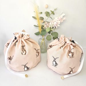 May include: Two drawstring bags with a pale pink fabric, patterned with brown and black rabbits. Each bag has a white ribbon drawstring and a small wooden bead. A vase of flowers is in the background.