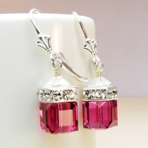 Könnte beinhalten: Ein Paar silberne Ohrhänger mit einem rosa Kristallwürfel und einem klaren Kristallhalo. Die Ohrringe haben einen silbernen Hakenverschluss.
