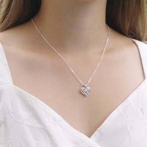 Puede incluir: Un collar plateado con un colgante en forma de coraz&oacute;n. El colgante presenta un dise&ntilde;o calado delicado. El collar se lleva sobre una parte superior blanca. La cadena es fina y el colgante est&aacute; centrado.