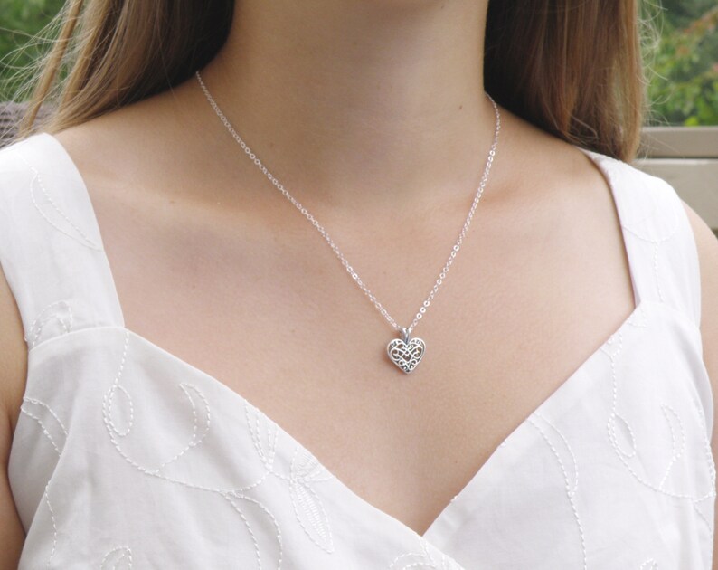 Puede incluir: Un collar plateado con un colgante en forma de coraz&oacute;n. El colgante presenta un dise&ntilde;o calado delicado. El collar se lleva sobre una parte superior blanca. La cadena es fina y el colgante est&aacute; centrado.