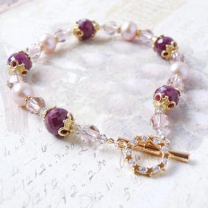 May include: A delicate bracelet featuring alternating burgundy and pale pink beads, interspersed with clear and gold-toned accents. The bracelet is secured with a gold-toned clasp with a decorative starburst design.
