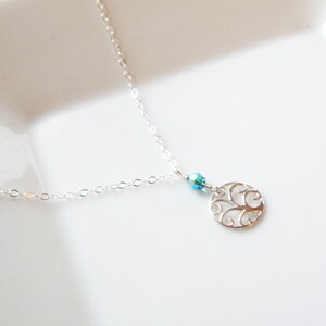 Puede incluir: Un delicado collar de plata con un peque&ntilde;o colgante circular calado. El colgante presenta un dise&ntilde;o intrincado, y una peque&ntilde;a cuenta azul iridiscente lo acompa&ntilde;a. El collar descansa sobre una superficie blanca.