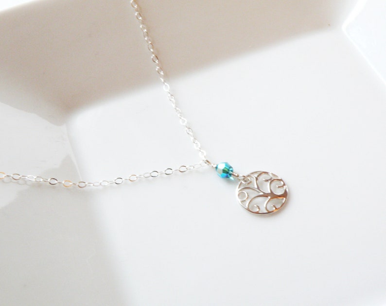 Puede incluir: Un delicado collar de plata con un peque&ntilde;o colgante circular calado. El colgante presenta un dise&ntilde;o intrincado, y una peque&ntilde;a cuenta azul iridiscente lo acompa&ntilde;a. El collar descansa sobre una superficie blanca.