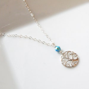 Puede incluir: Un delicado collar de plata con un colgante circular calado y una peque&ntilde;a cuenta azul iridiscente. La fina cadena es elegante. El colgante presenta un dise&ntilde;o decorativo con volutas.