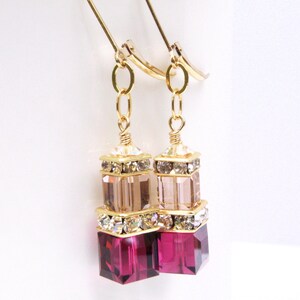 May include: Gold dangle earrings with three stacked crystal beads. The top bead is a light pink color, the middle bead is clear, and the bottom bead is a deep magenta color.