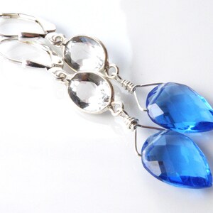 May include: A pair of silver-toned drop earrings. Each earring features a clear, oval-shaped stone above a faceted, teardrop-shaped blue gemstone. The earrings have lever-back closures and are displayed on a white background.