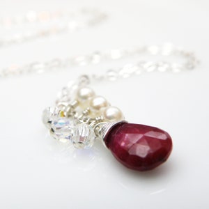 May include: A silver chain necklace with a red gemstone pendant. The pendant is surrounded by small white pearls and clear crystals.