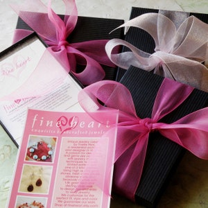 May include: A pink and black gift box with a pink ribbon and a card that reads "fine heart exquisite handcrafted jewelry". The card has a picture of a heart-shaped necklace with turquoise stones and a pair of earrings with turquoise stones.