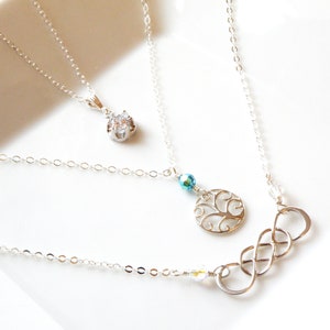 Puede incluir: Una colecci&oacute;n de collares de plata con varios dise&ntilde;os de colgantes. Un collar presenta una gema transparente, otro un &aacute;rbol de la vida calado y el tercero un s&iacute;mbolo de infinito. Los collares se muestran sobre una superficie blanca.