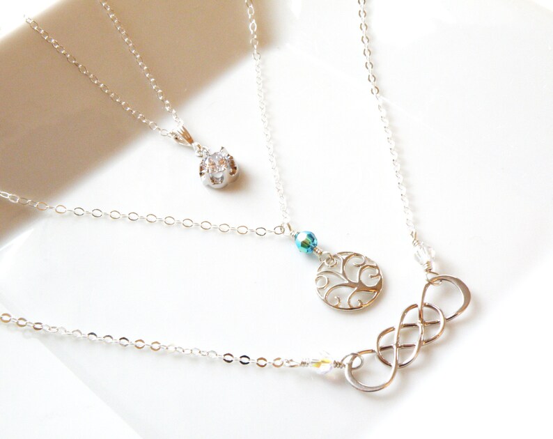 Puede incluir: Una colecci&oacute;n de collares de plata con varios dise&ntilde;os de colgantes. Un collar presenta una gema transparente, otro un &aacute;rbol de la vida calado y el tercero un s&iacute;mbolo de infinito. Los collares se muestran sobre una superficie blanca.