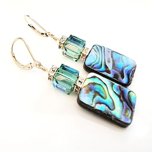 May include: A pair of dangle earrings featuring rectangular abalone shell pendants with iridescent blue and green hues. Each earring has a teal crystal cube and is accented with small, clear crystals and gold-colored metal hardware.