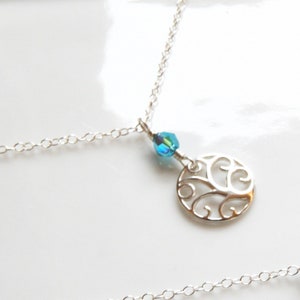 Puede incluir: Un collar de plata con una cadena delicada y un colgante circular. El colgante presenta un dise&ntilde;o calado y est&aacute; adornado con una peque&ntilde;a cuenta azul iridiscente. El collar se muestra sobre una superficie blanca.