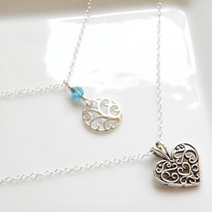 Puede incluir: Dos collares de plata con cadenas finas. Uno presenta un colgante de coraz&oacute;n calado, el otro un colgante circular con un dise&ntilde;o de &aacute;rbol y una cuenta de cristal azul. Los collares se exhiben sobre una superficie blanca.