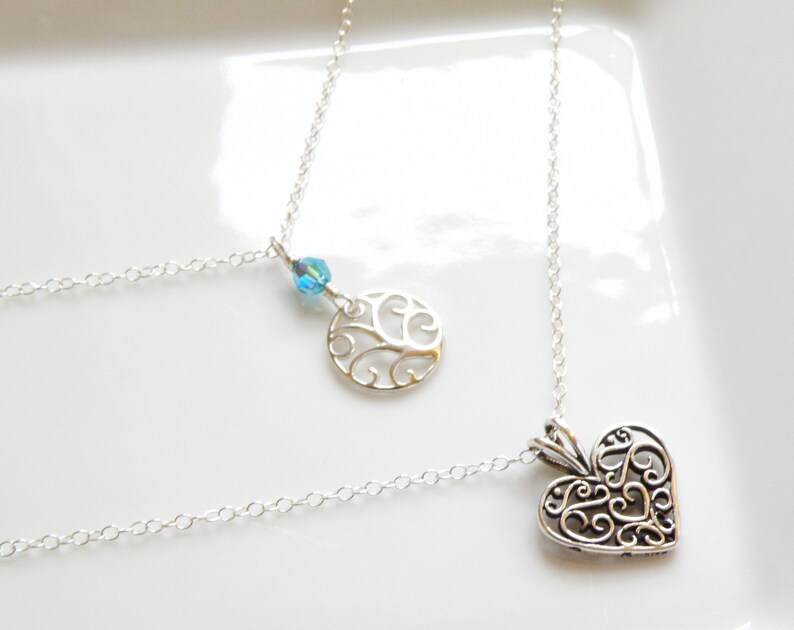 Puede incluir: Dos collares de plata con cadenas finas. Uno presenta un colgante de coraz&oacute;n calado, el otro un colgante circular con un dise&ntilde;o de &aacute;rbol y una cuenta de cristal azul. Los collares se exhiben sobre una superficie blanca.