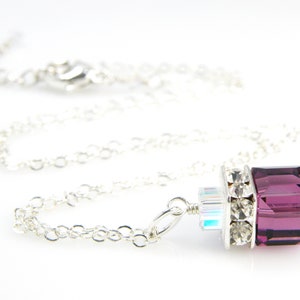 May include: A silver chain necklace with a pendant featuring a large, square, purple crystal surrounded by smaller clear crystals.